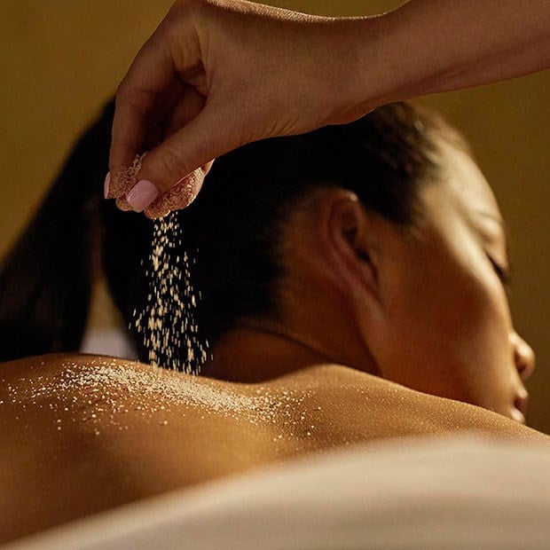 A lady lies on a massage table, a gentle hand dusting powder on her shoulder.
