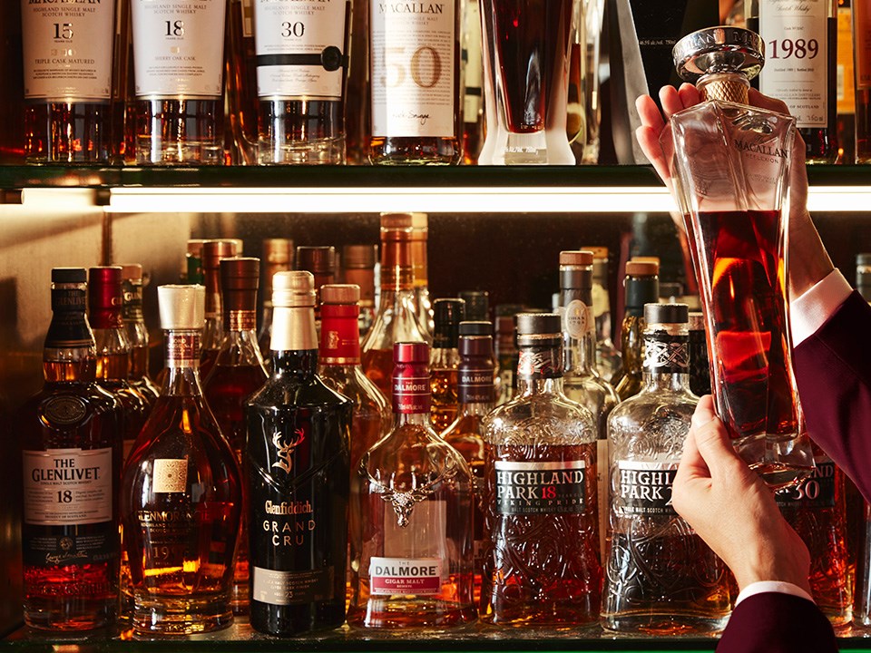 Various whiskey bottles on a glass shelf. The arms of a bartender are shown holding up a bottle.