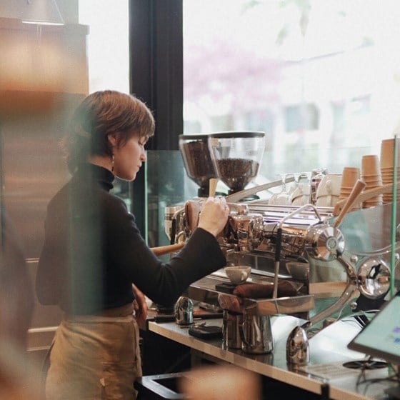 At the café a female barista pours a shot of expresso using the large silver coffee machine