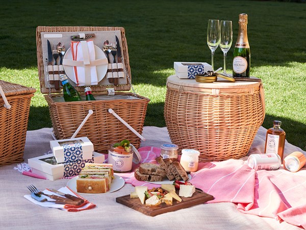 Picnic food laid out in front of wicker basket with glasses of champagne, fresh produce and light snacks on a blanket