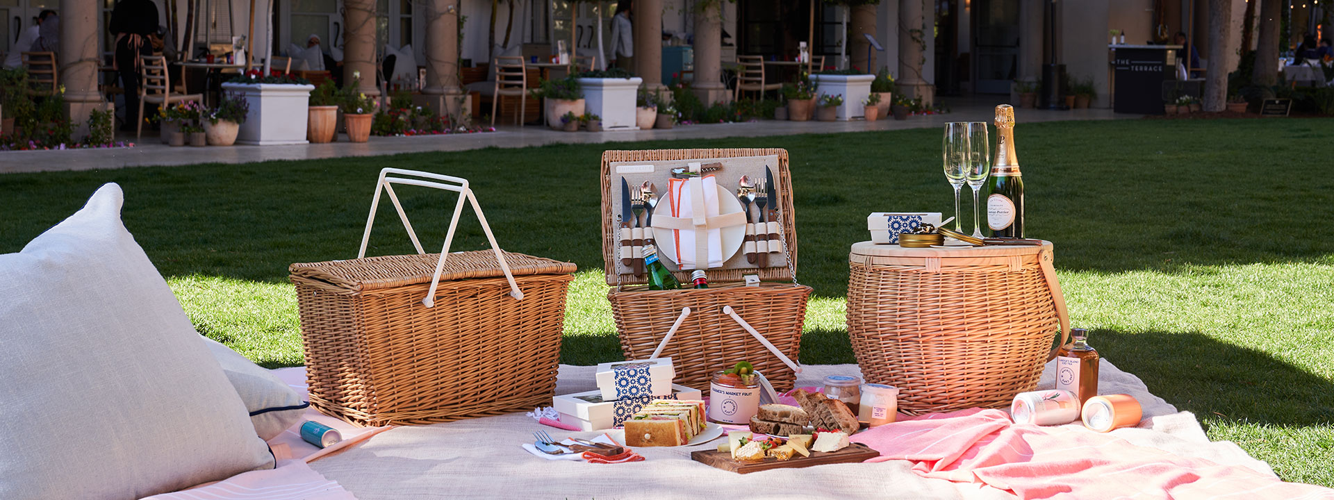 Picnic food laid out in front of wicker basket with glasses of champagne, fresh produce and light snacks on a blanket