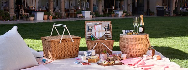 Picnic food laid out in front of wicker basket with glasses of champagne, fresh produce and light snacks on a blanket