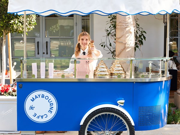 Woman in a peach colored apron holding a gelato cone standing behind a blue and white gelato cart outside
