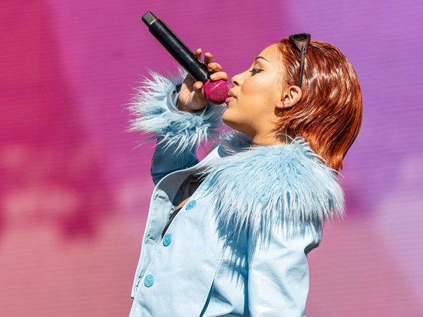 Doja Cat singing with a pink microphone in front of a large pink background on a digital screen.