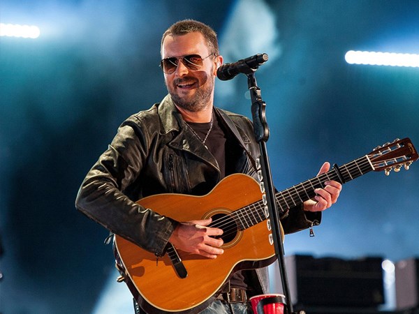 Eric Church wearing sunglasses holding an acoustic guitar and singing on stage.,