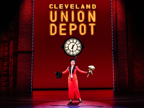 A woman wearing a red dress and black hat on a theater stage in front of a large clock. Above her hangs a sign with lights that says: Cleveland union depot.
