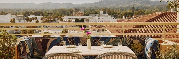 An outside table with a pink flower in a vase at a restaurant overlooking a city skyline with rolling hills and buildings.