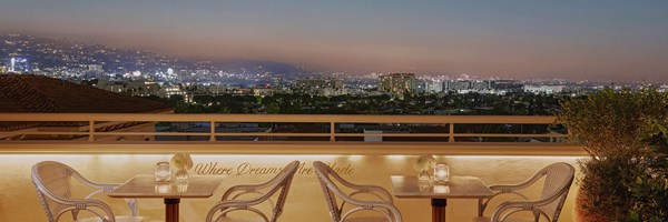 Two well lit tables in an outside dining area at sunset. City lights can be seen on rolling hills in the background.