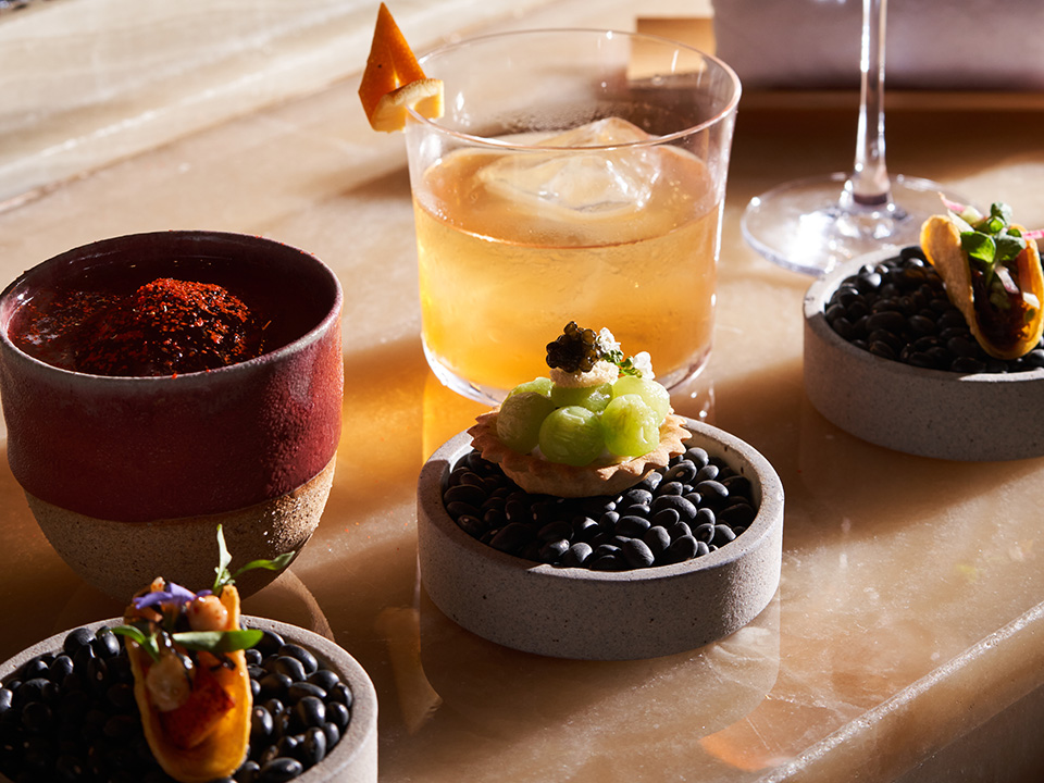 Two cocktails, one garnished with an orange peel, and three small bite sized appetizers sitting on stone plates with black pebbles.