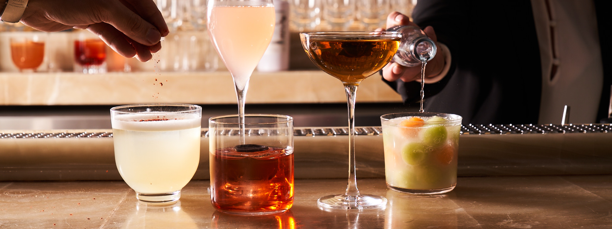 Five cocktails lined up on a bar with two hands in the shot pouring and garnishing