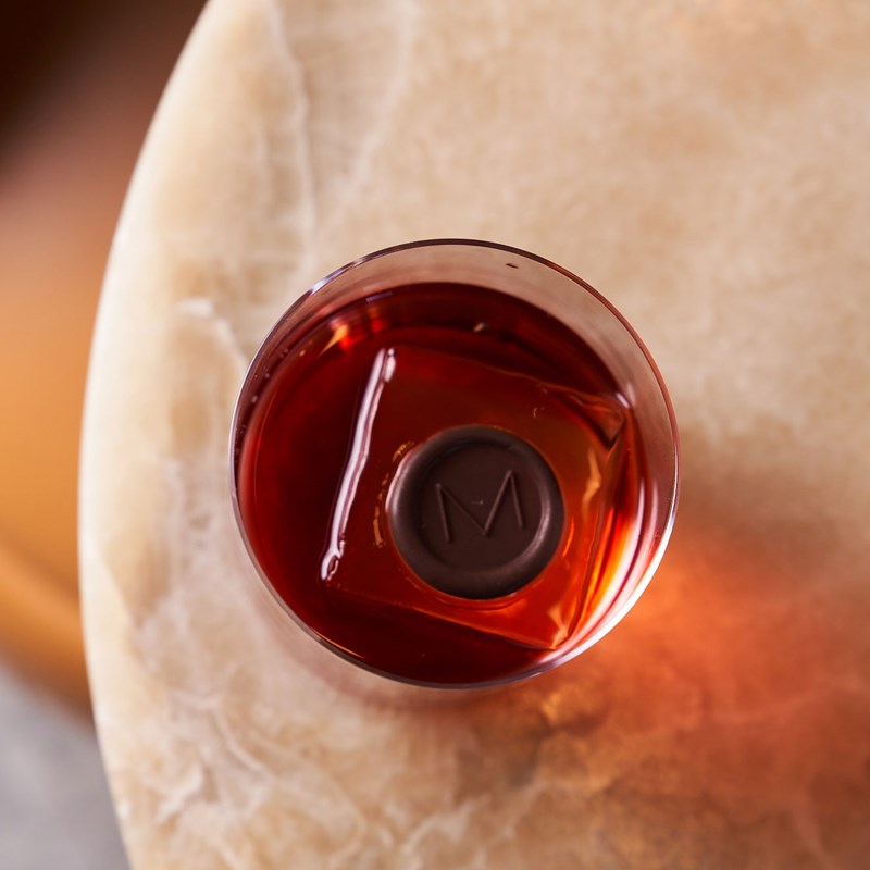 Overhead shot of a cocktail with a square ice cube and chocolate medallion stamped with an M as a garnish