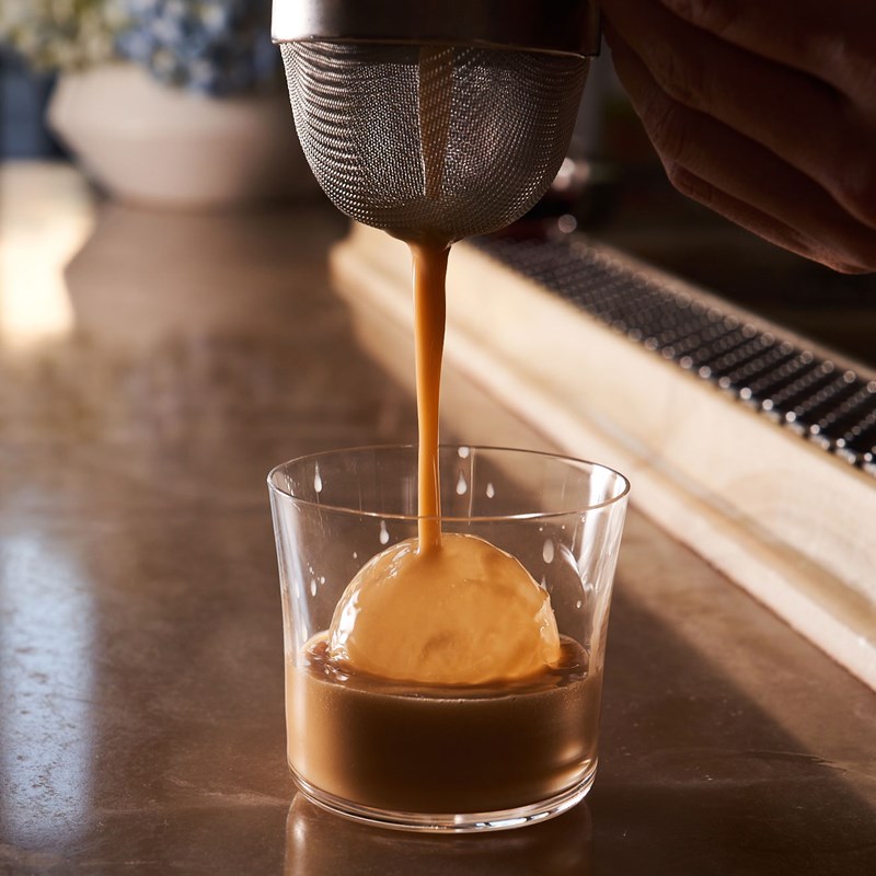 Cocktail being poured from a strainer into a glass over a spherical cube