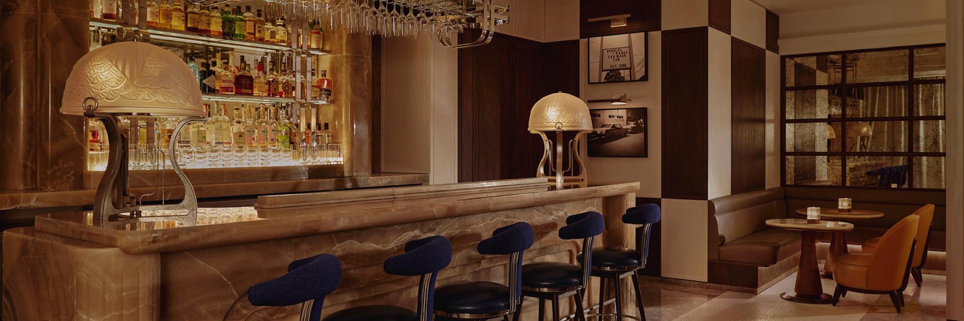 A marble bar with five blue stools in a dimly lit, elegant bar. Bottles of spirits line the glass shelves behind the bar.