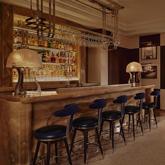 A marble bar with five blue stools in a dimly lit, elegant bar. Bottles of spirits line the glass shelves behind the bar.