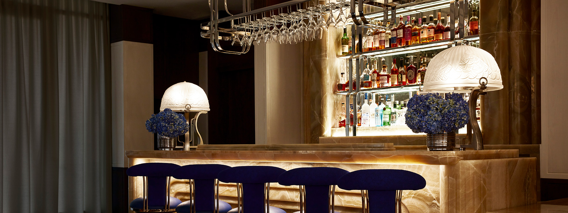 Five dark blue stools at The Maybourne Bar. Behind the bar are shelves of various alcohol brands on display.