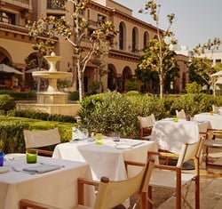 Outside dining tables and chairs with white umbrellas. Behind, there's a fountain in a garden surrounded by bushes and tree.