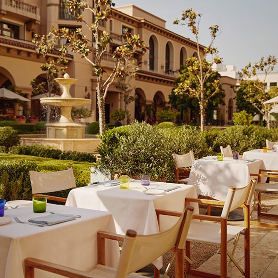 Outside dining tables and chairs with white umbrellas. Behind, there's a fountain in a garden surrounded by bushes and tree.