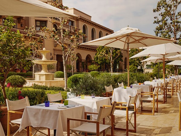 Outside dining tables and chairs with white umbrellas. Behind, there's a fountain in a garden surrounded by bushes and tree.