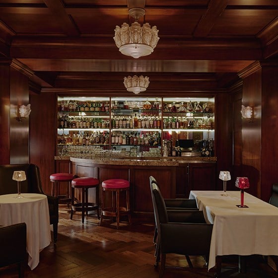 A dimly lit marble bar with red stools. Whisky brands line the glass shelves behind the bar. There are tables with lamps.