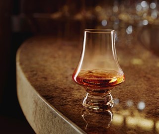 A glass filled with a small shot of whisky on a brown, marble bar.