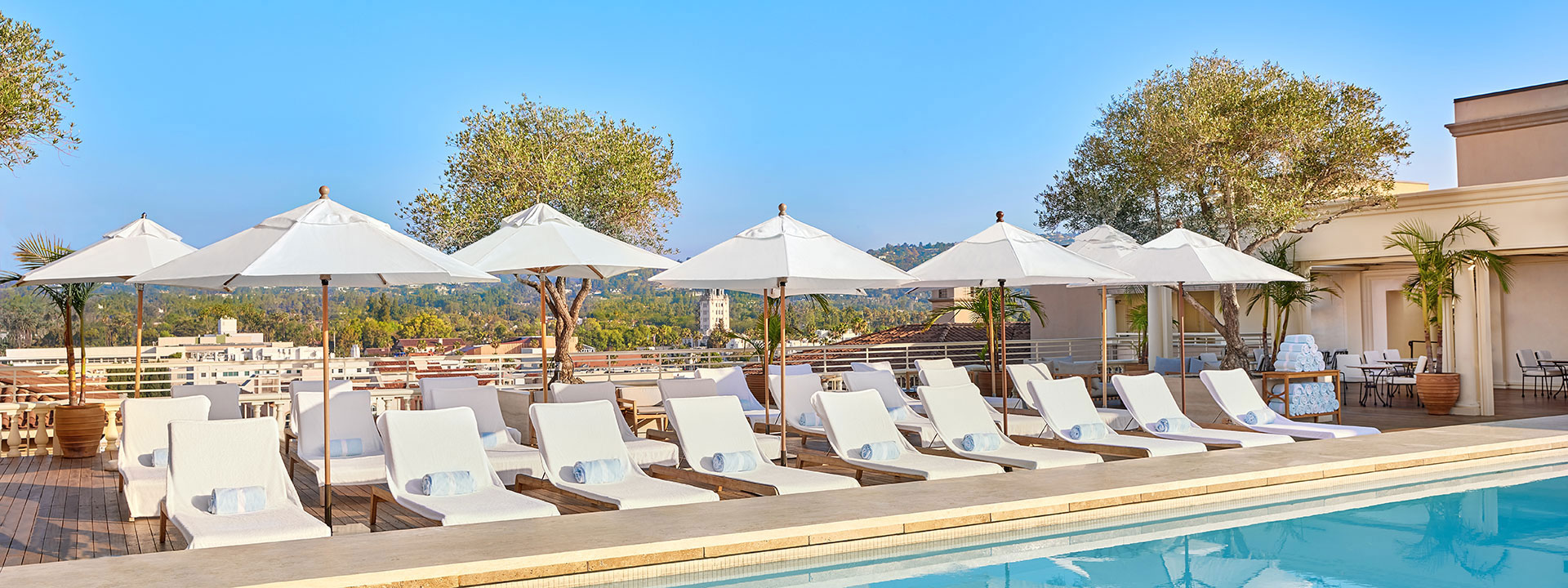 Rows of white sunbeds with parasols in front of the rooftop pool with a view over the L.A skyline