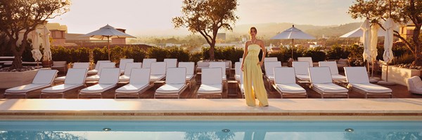 A female model wearing a yellow jumper in front of white sunbeds by a rooftop pool.