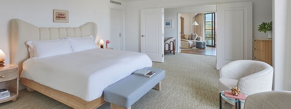 A king sized bed with a white comforter in a hotel suite. In the distance is a living space with a curved beige couch.