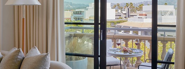 A sliding glass door to a balcony in a hotel. There is a garden table with two wine glasses and palm tree and buildings in the distance.
