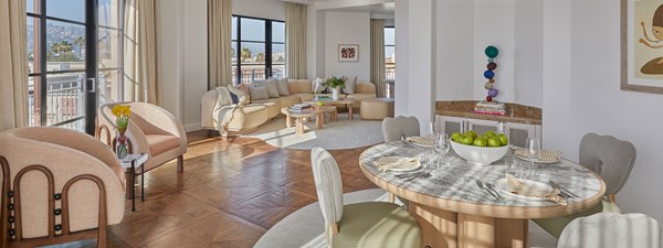 The dining area of a hotel room. There's a bowl of green apples on the table. The living room is in the background with a beige couch.