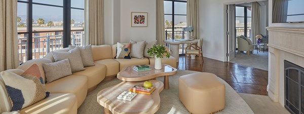 A curved beige couch in the living area of a hotel suite. There are large windows letting in sunlight and a fireplace.