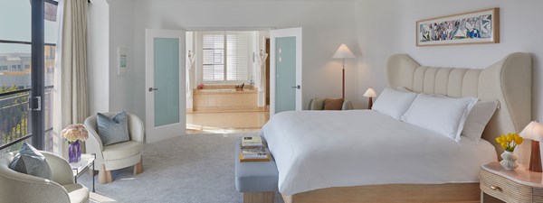 A king sized bed with a white comforter in the bedroom of a hotel suite. Doors are open in the background leading to a bathtub.