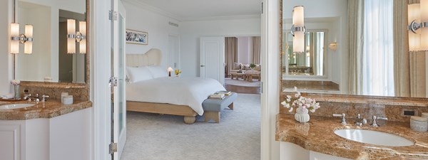 A view from the bathroom of a hotel suite into the bedroom. There are two vanity sinks with mirrors and a king sized bed white sheets.