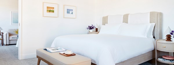 A king sized bed with a white comforter in a hotel. There are two nightstands by the bed with vases willed with purple flowers.