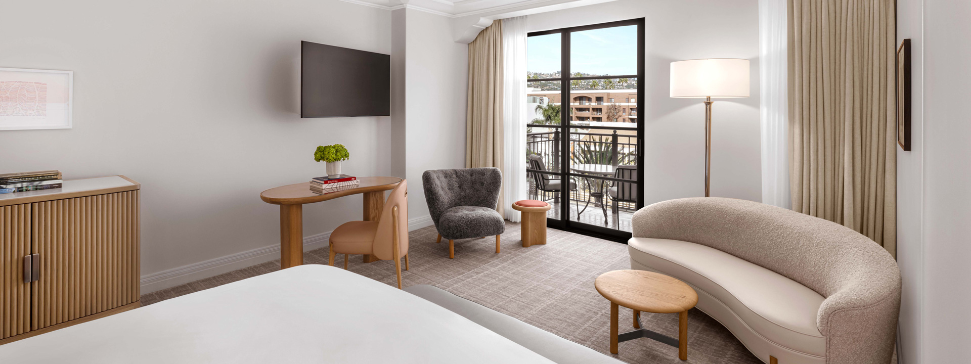 The edge of a king sized bed in a hotel room. There's a curved ivory couch and a garden table and chairs outside on the balcony.