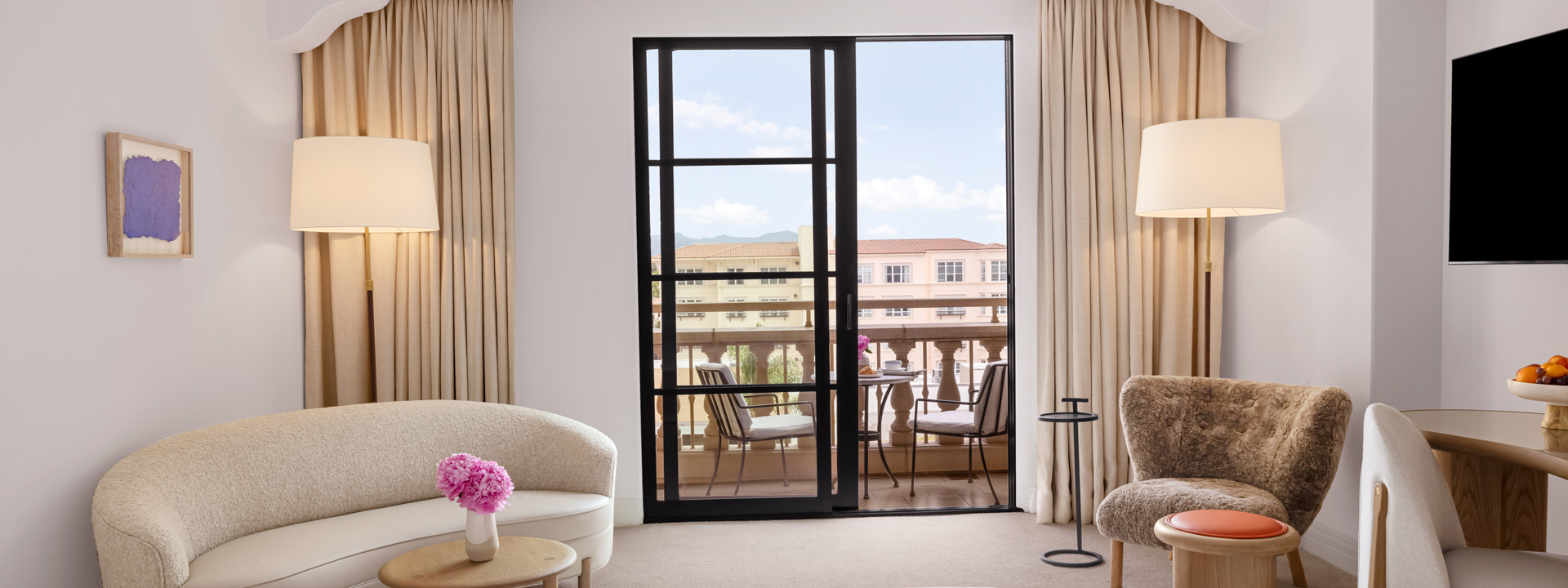 The living area of a hotel room. There is a white bean shaped couch, a wall mounted tv, and a table and chairs outside on the balcony.