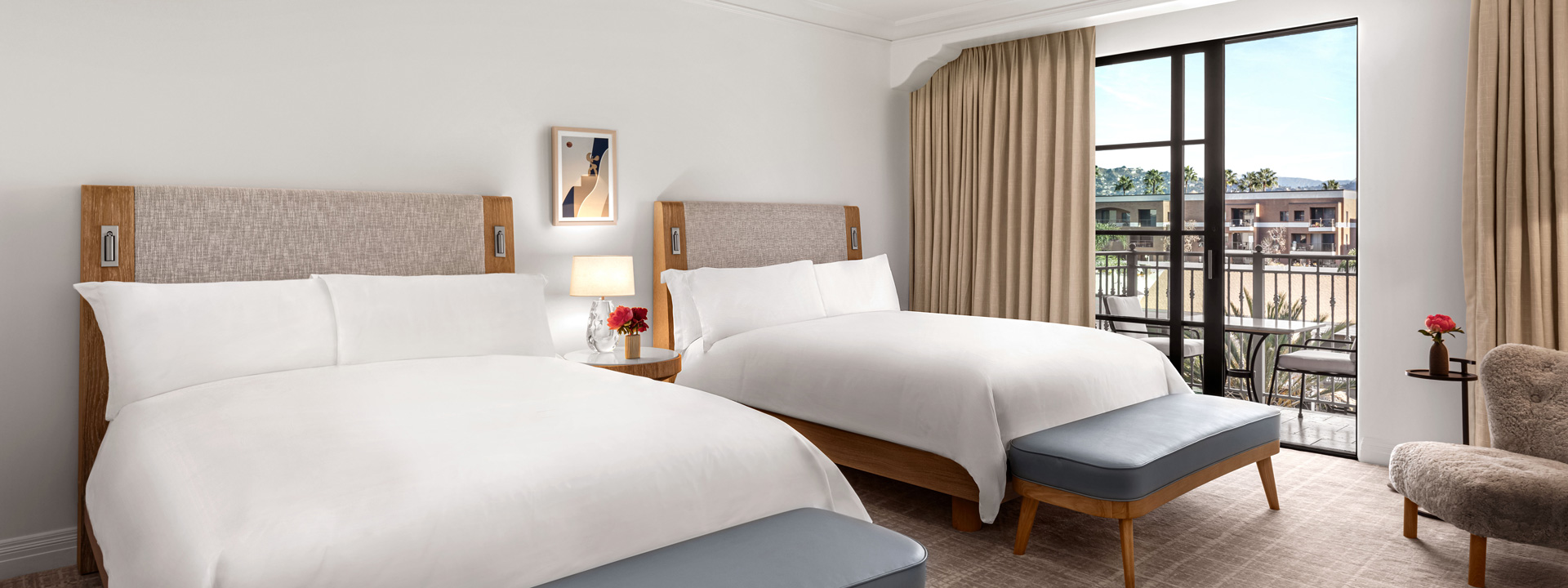 Two queen sized beds in a hotel room at the Maybourne Beverly Hills. A table and two chairs can be seen out on a balcony.