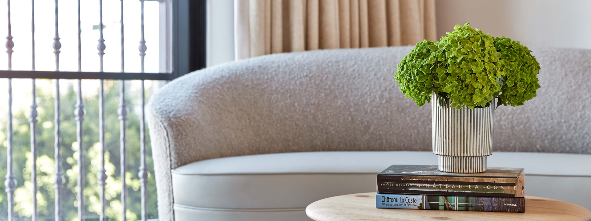 A curved ivory couch and a small wooden table. On the table is a vase of green hydrangeas sitting on top of two books.