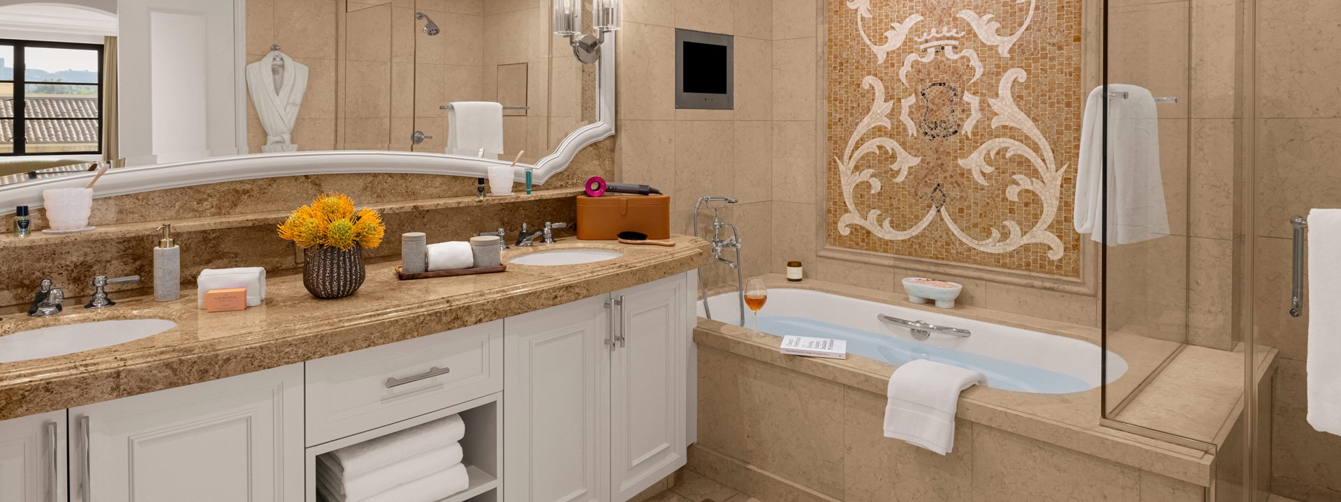 Two sinks and a bathtub in a brown hotel bathroom. There is a large mosaic on the wall.