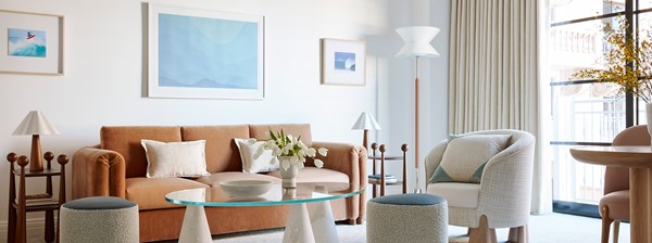 The living area of the Rodeo Suite at the Maybourne Beverly Hills hotel. There is a brown couch, a white chair, and a glass table.