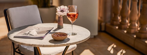 A marble garden table on a private terrace. There are nuts on a plate, a magazine, a glass of wine, and a pink flower in a vase.