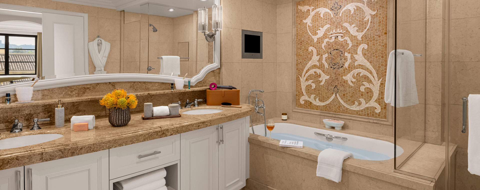 The bathroom of a hotel room with two sinks and a large bathtub. There is a mosaic wall above the bath tub.