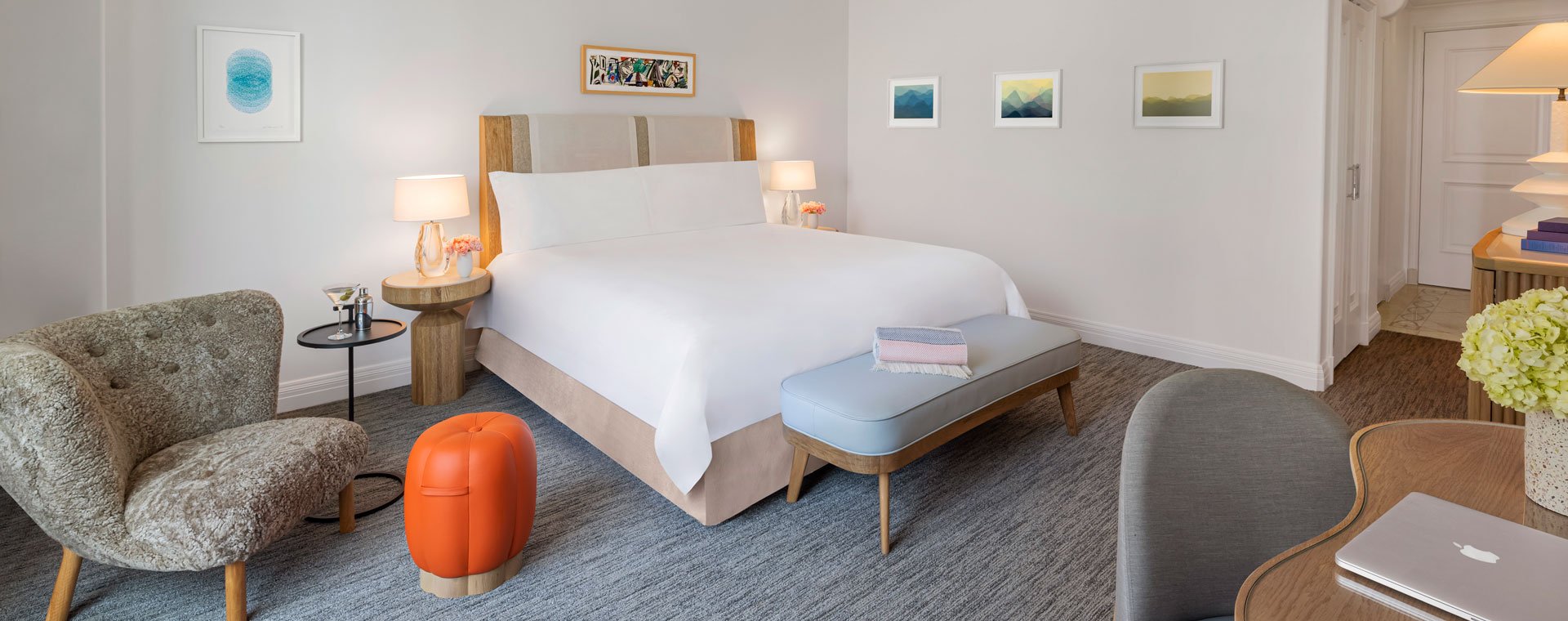 A king sized bed in a hotel room. There is an orange pumpkin shaped stool and a martini glass and shaker on a side table.