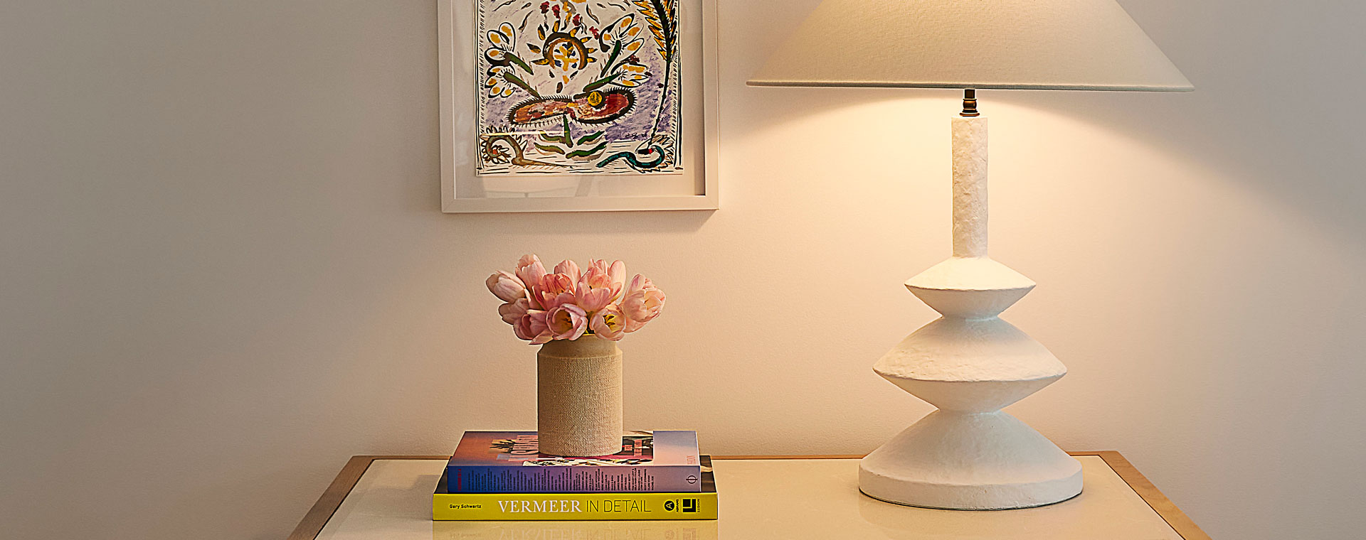 A table being lit by a lamp. There is a vase of pink flowers sitting on top of two books and a modern painting on the wall.
