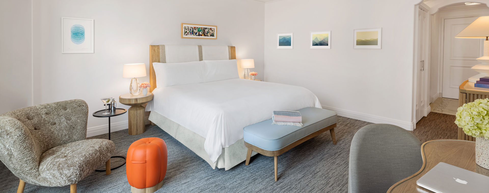A king sized bed in a hotel room. There is a small grey chair with an orange, pumpkin shaped foot stool.