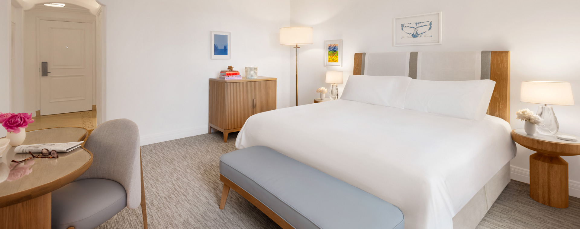 A king sized bed in a hotel room. There is a wooden table with a vase of purple flowers, a newspaper, and some reading glasses.