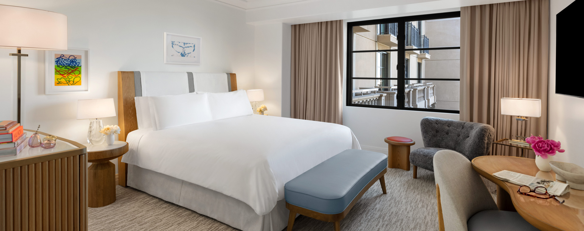 A king sized bed in a hotel room. There is a wall mounted TV and a large window letting in sunlight.