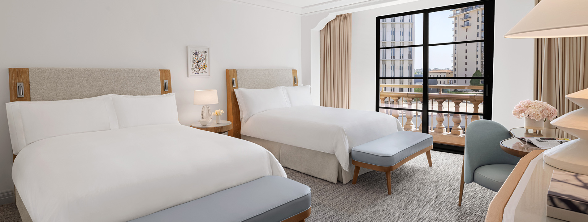 Two queen sized beds in a hotel room at the Maybourne Beverly Hills. A large window lets in light and tall buildings can be seen outside.