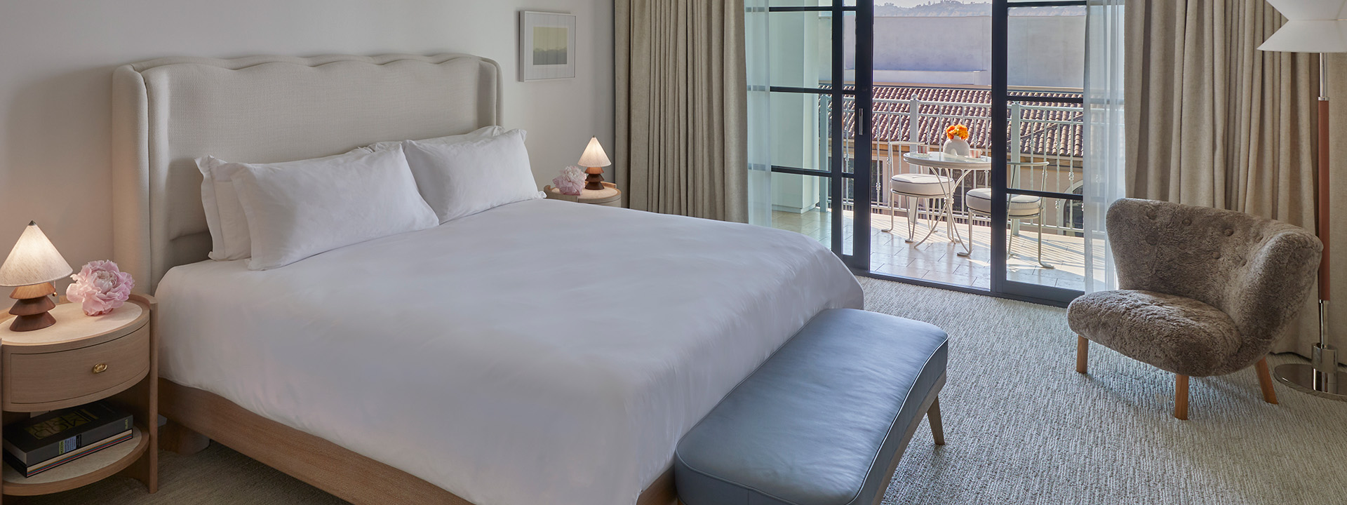 A king sized bed in a hotel room. There is a garden table and two chairs outside on a private terrace overlooking buildings.