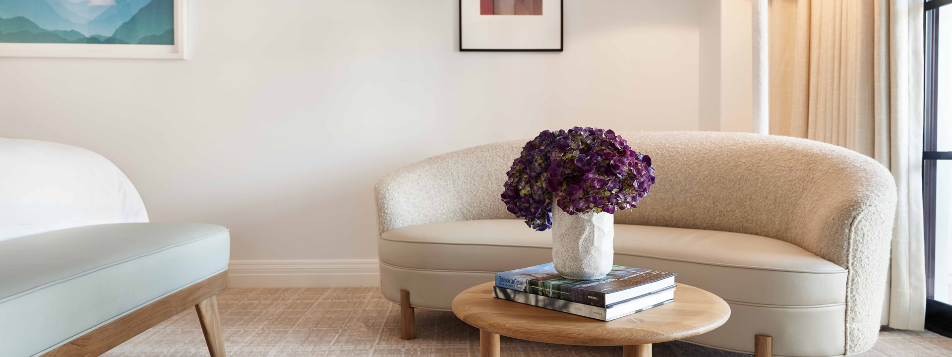 A white bean shaped couch and a light blue bench in the living area of a hotel room. A vase of purple flowers sites on a small wooden table.