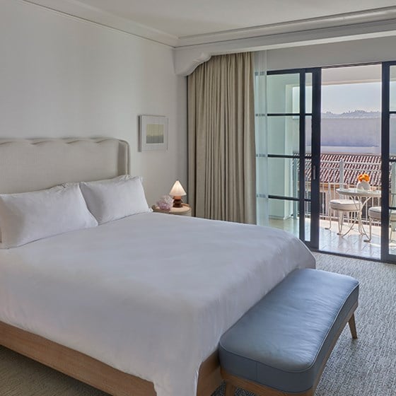 A king sized bed in a hotel room at the Maybourne Beverly Hills. There is a table and chair outside on the private terrace.